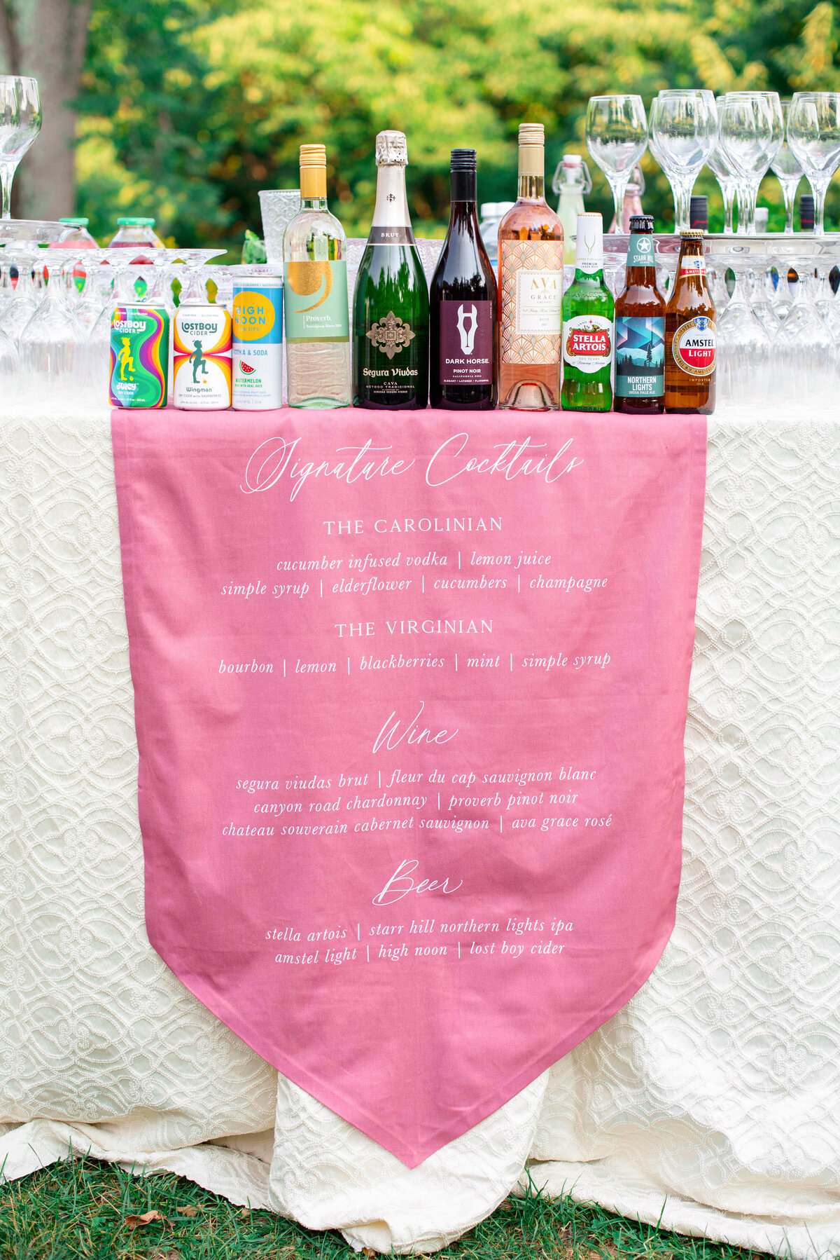 A table display with various liquor bottles, mixers, and beer options is set up. A pink cloth hangs in front, listing signature cocktails like The Carolinian and The Virginian, along with wine and beer selections. Glasses are arranged on top.