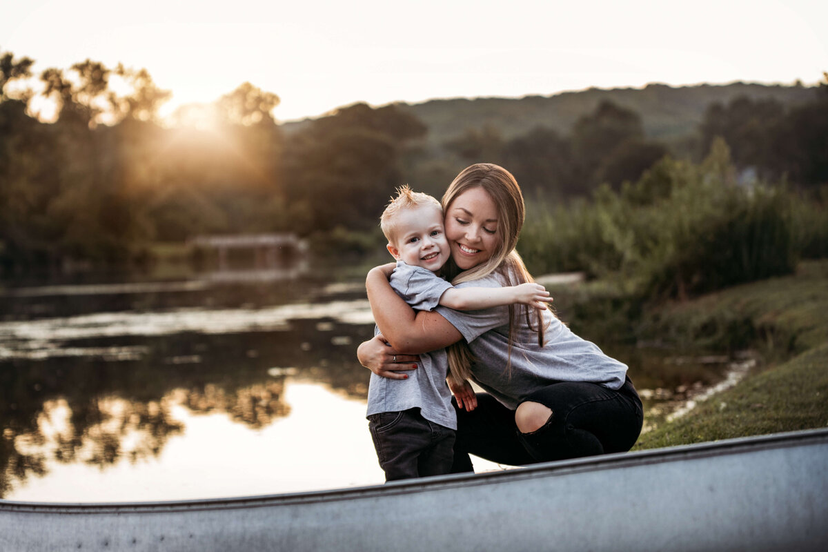 big sister and little brother hugging eachother behind a row boat in front of a lake at sunset
