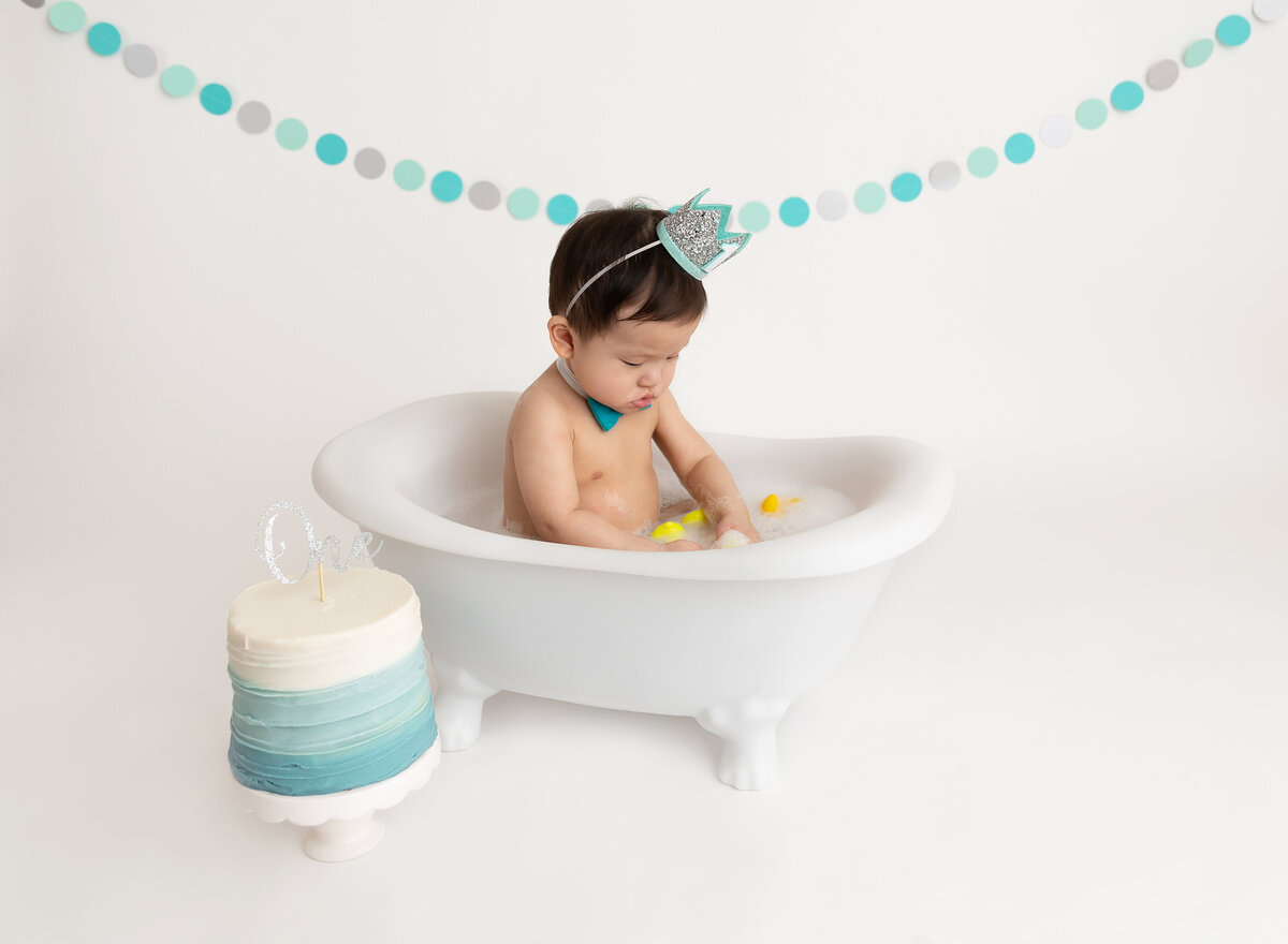 Baby boy sits in bathtub cleaning up from cake smash. Blue ombre cake is sitting in front of the tub.