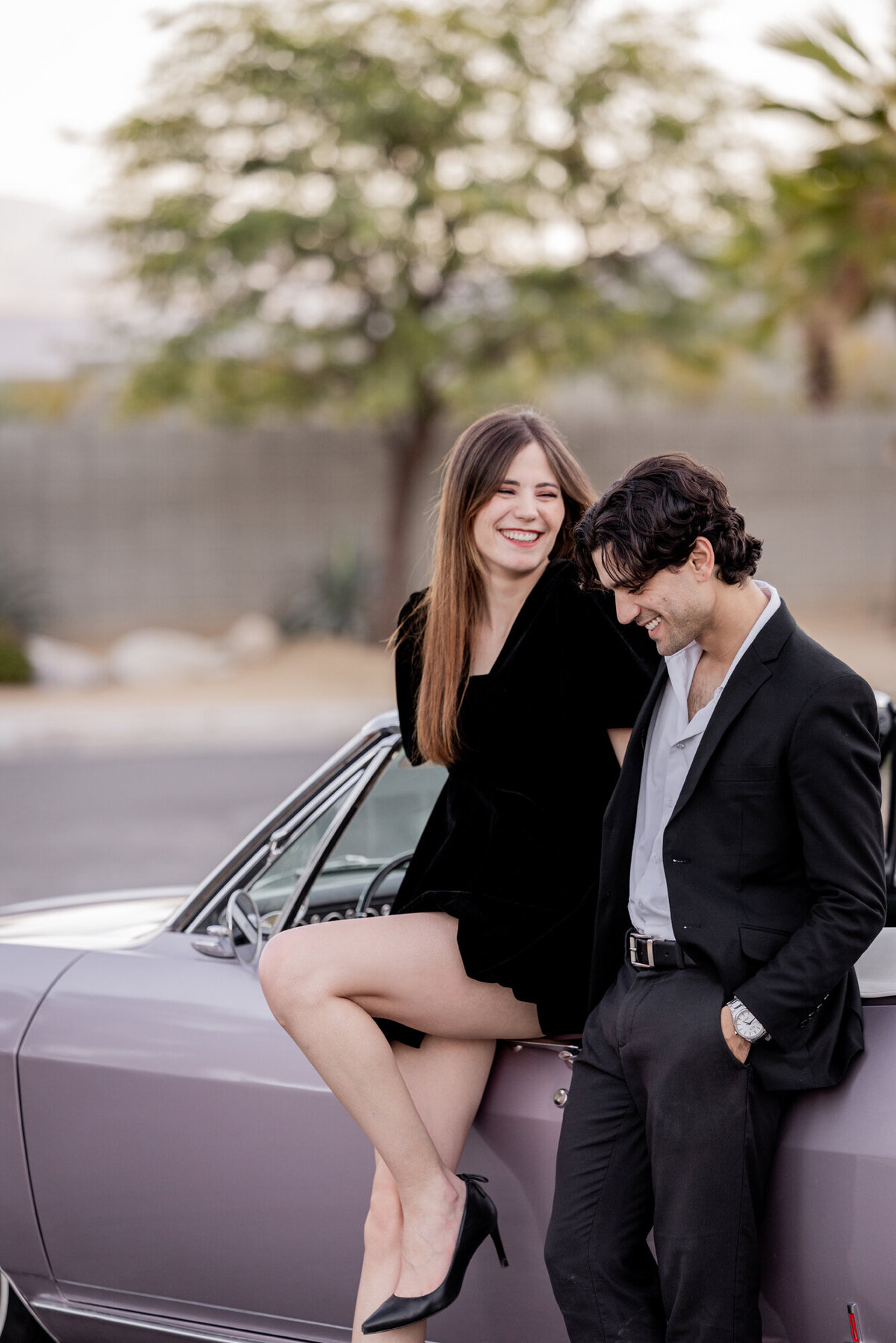 Couple sharing a light moment sitting on the hood of a classic car, dressed in modern black attire, with desert tones behind them.