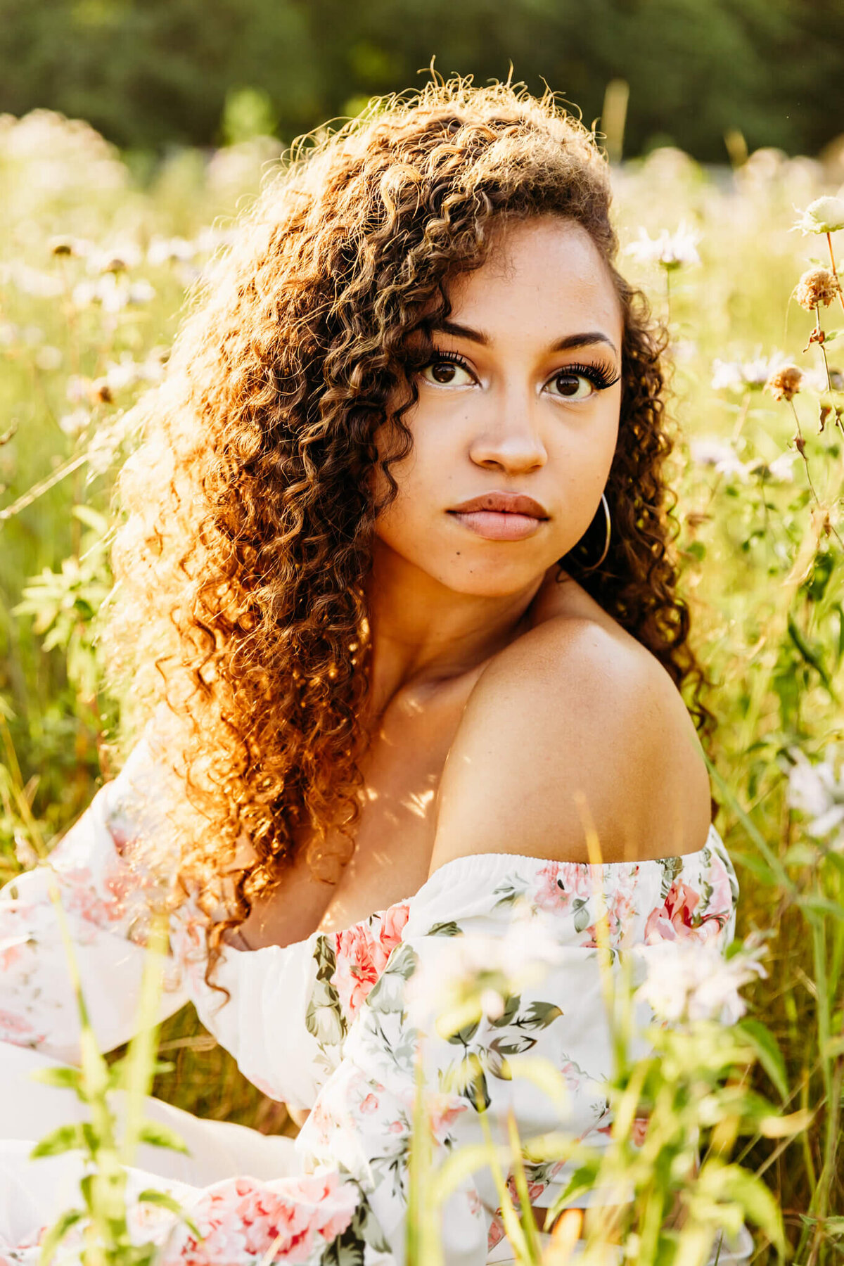 gorgeous high school senior girl looking over shoulder as she sits in a wildflower field for her senior photography session