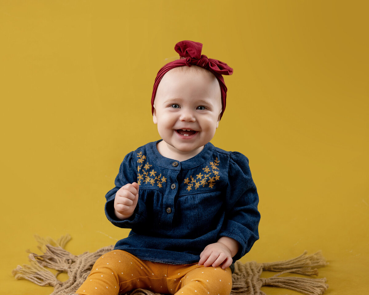 A happy girl sits on a yellow background while she wears a blue top and her Iowa Milestone Photographer takes her photos