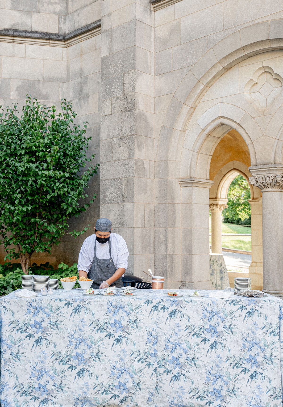 A chef wearing a mask and apron prepares food at an outdoor cooking station covered with a floral tablecloth. The setup is in front of a stone building with archways and a lush green tree to the left. The atmosphere appears calm and sunny.