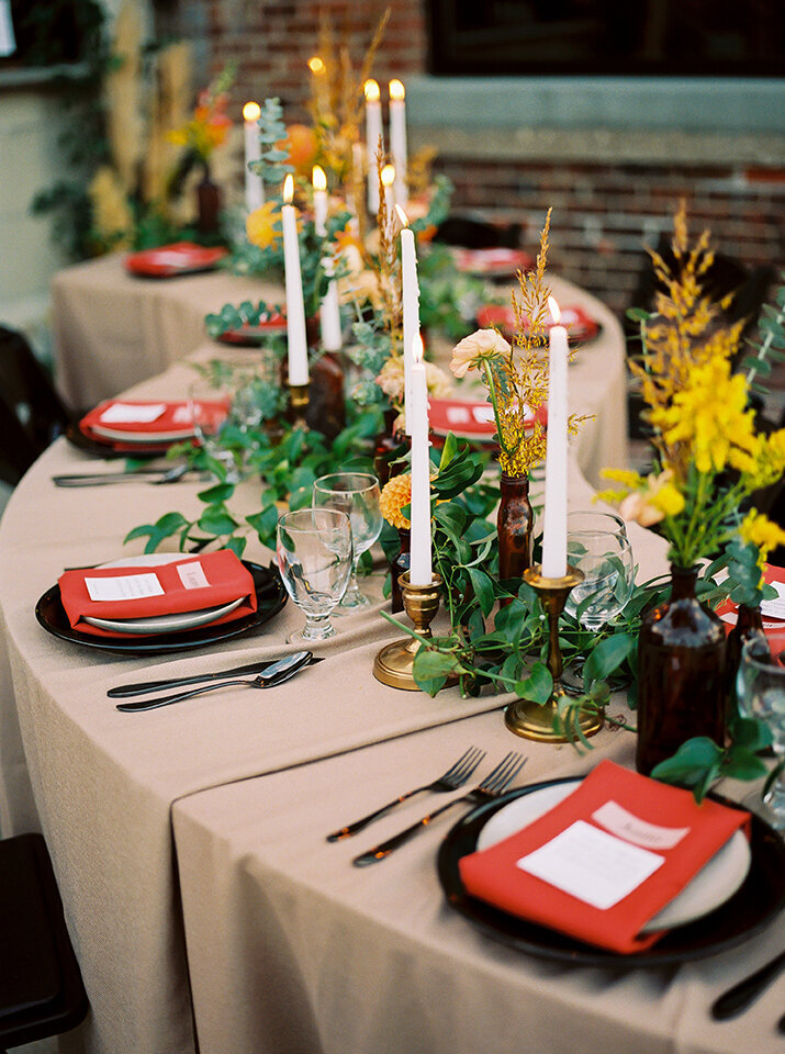 Curved table set with ivory table cloth, red napkins, candlesticks, and amber glassware with flowers.