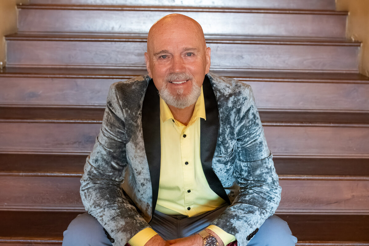 Portrait of a smiling man sitting on wooden steps wearing a patterned blazer and yellow shirt, photographed by Vyrl Photo for Tucson brand photography.