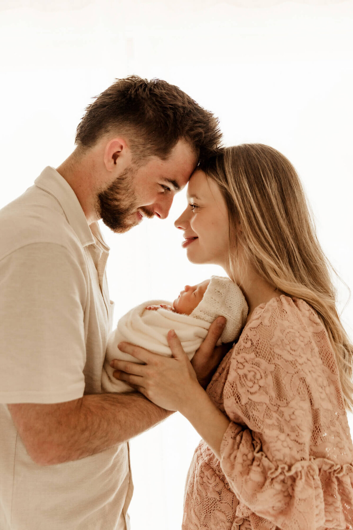 Mother and father touching foreheads with the light behind them as they hold their baby girl.
