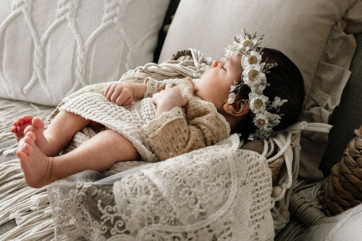 A beautiful portrait of a peaceful newborn baby girl wearing a charming floral and greenery crown, sleeping in a rustic basket with soft layers