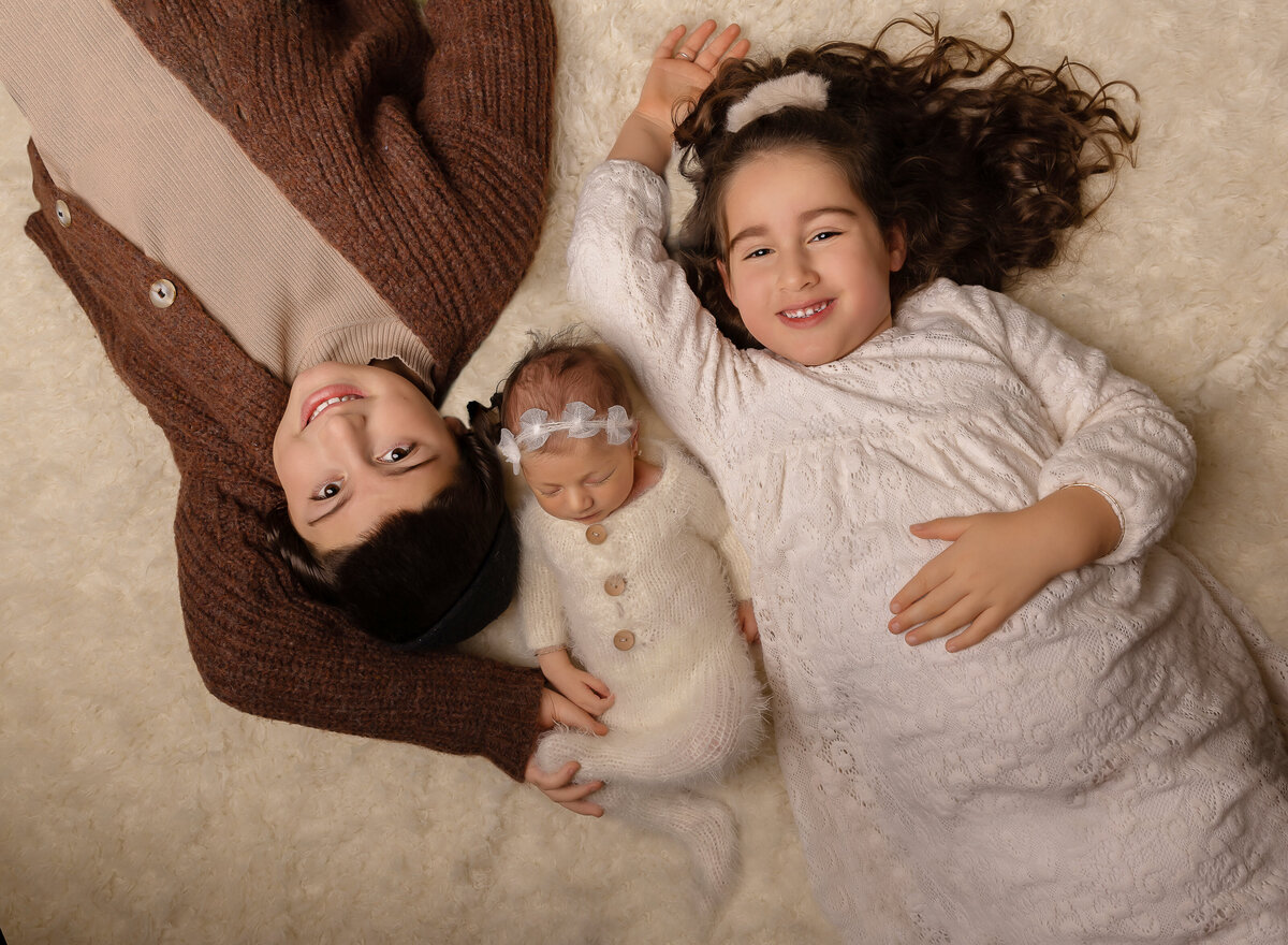 Aerial image. Brother and sister are laying atop of a beige fuzzy rug with their new baby sister laying between them. Big brother and sister are smiling at the camera. Captured by top Brooklyn newborn photographer Chaya Bornstein