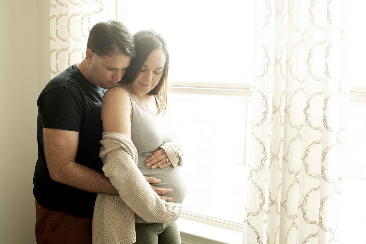 A serene maternity photo of mom and dad standing in front of a window, with soft natural light pouring in as mom lovingly cradles her baby bump and dad stands beside her with a gentle smile, captured by Chunky Monkey Photography.