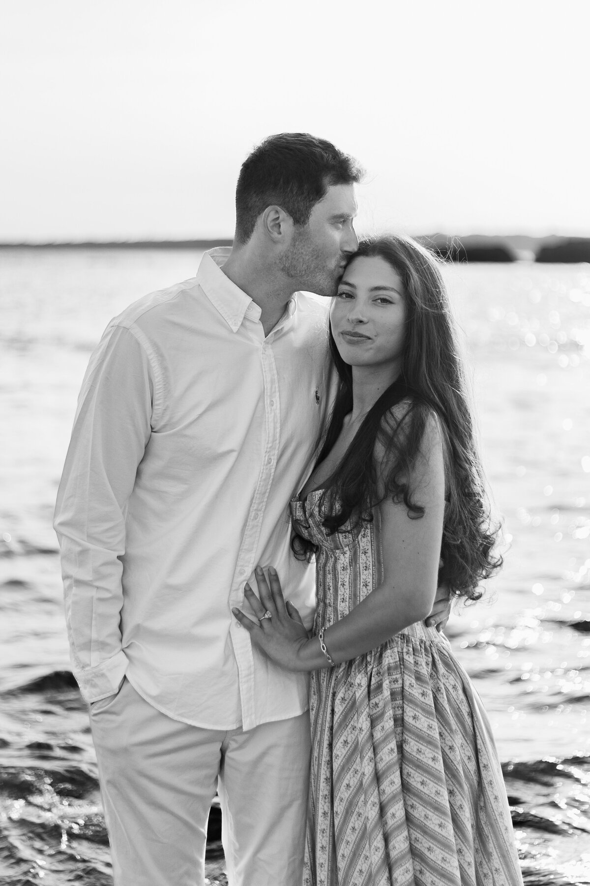Collins Beach Newport RI | Kelsey Sheehan Photography Timeless Rhode Island Weddings | A couple stands by a serene waterfront; the man in a light shirt kisses the woman's head. She wears a patterned dress, smiling softly. Romantic and peaceful.