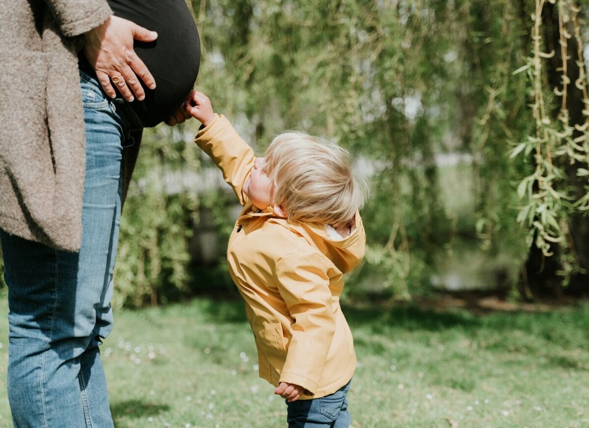 Young boy in yellow raincoat gently touching his mother’s pregnant belly outdoors.