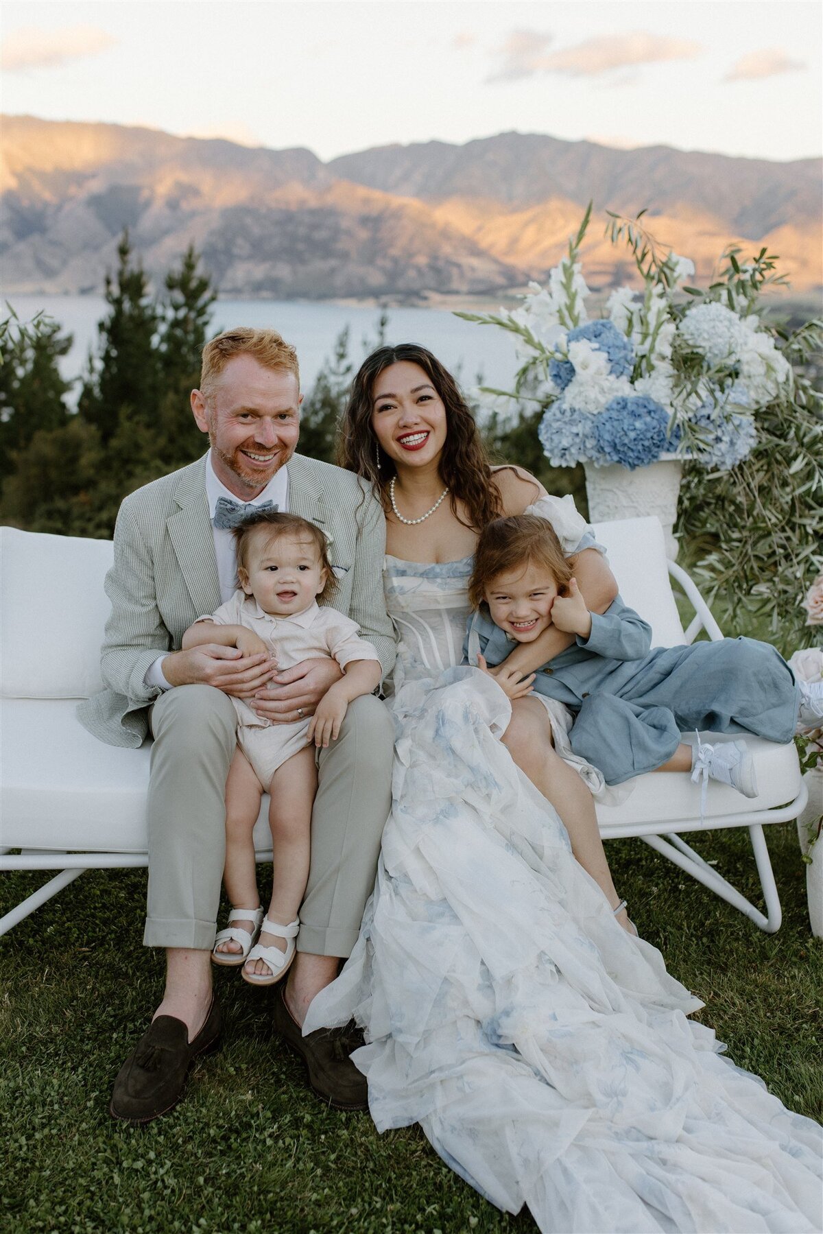 A bride and groom sit on a white outdoor couch with their two young children in their laps, surrounded by lush flowers and a stunning mountain and lake backdrop during golden hour.