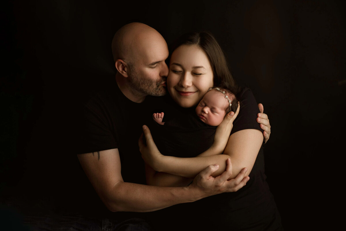 Parents cuddling newborn on dark backdrop - intimate newborn family portrait in Minneapolis