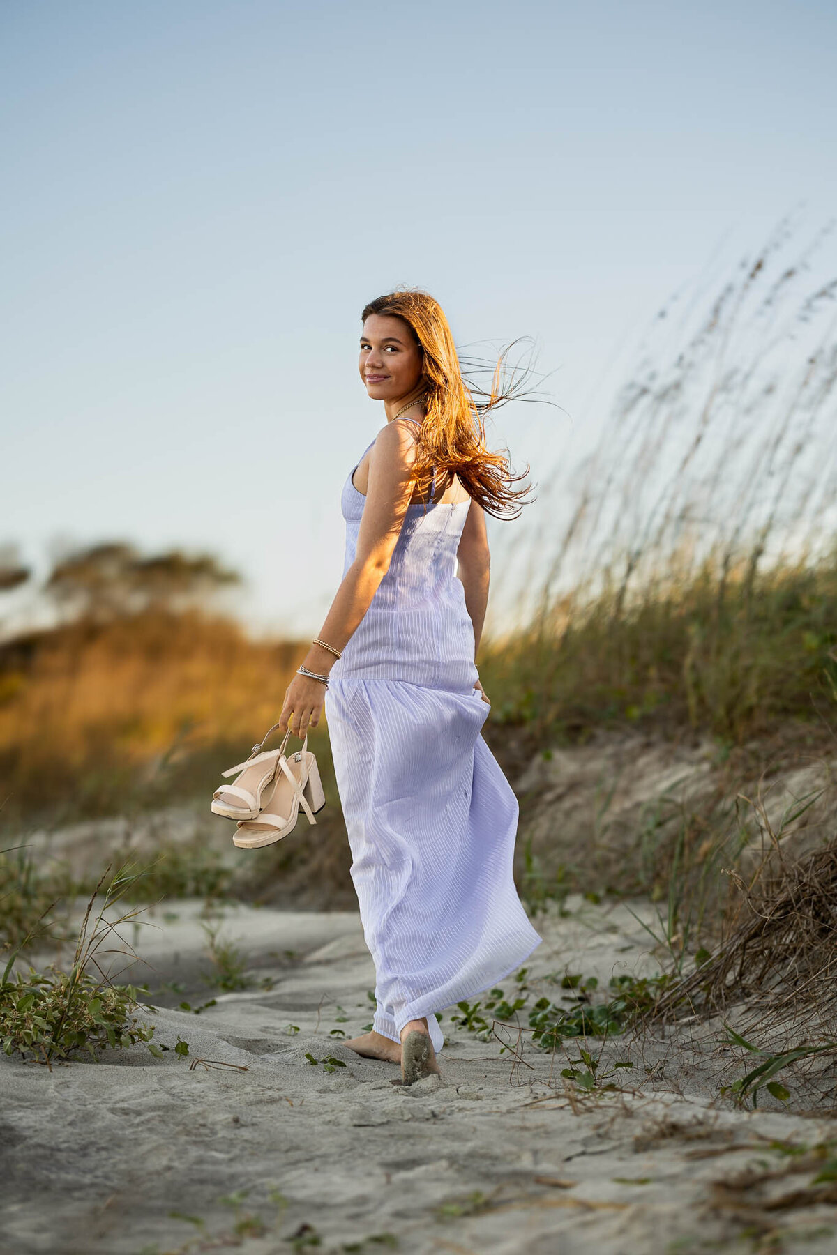 A high school senior girl poses gracefully in a lavender dress in an open field, looking over her shoulder with a soft and dreamy expression.