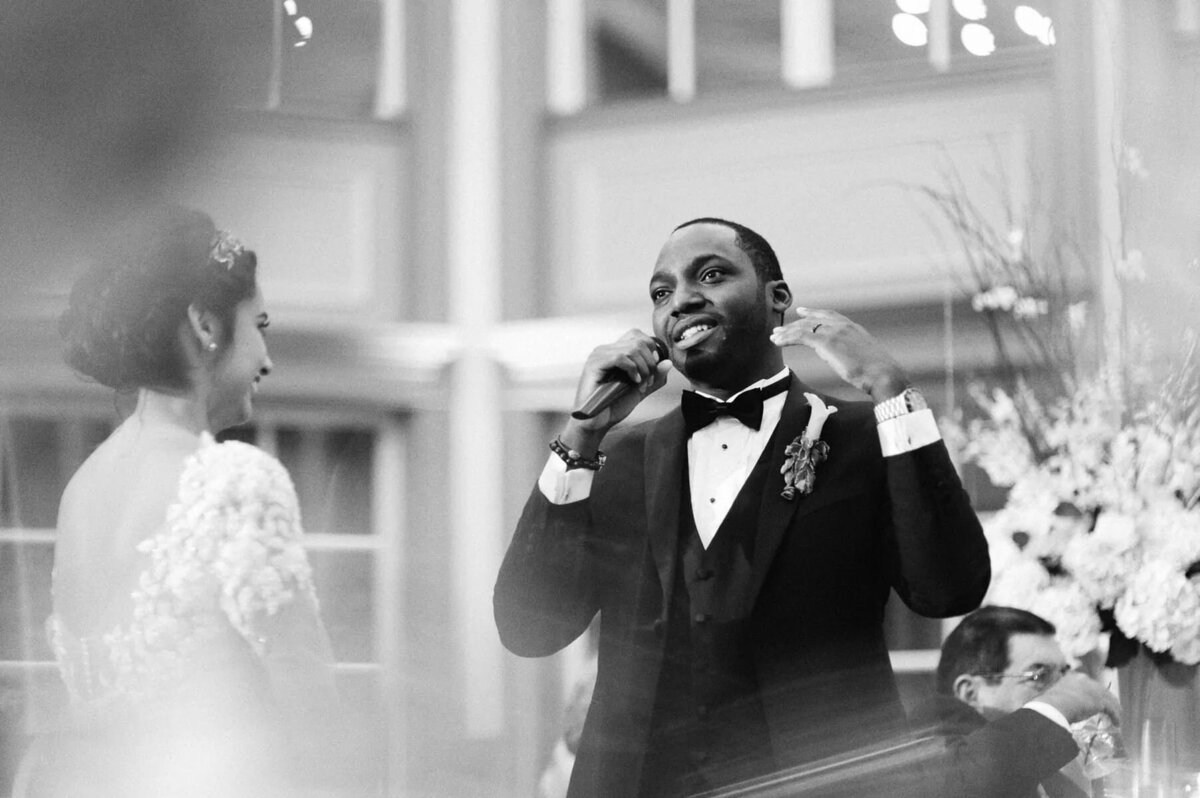A groom talking into a microphone with his bride in front of him.