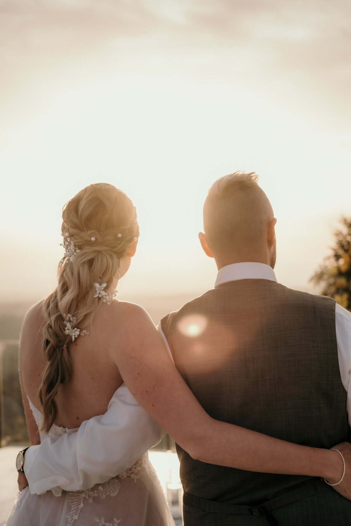 Von hinten betrachtet, sich im Arm haltend blickt das Hochzeitspaar in den Sonnenuntergang.