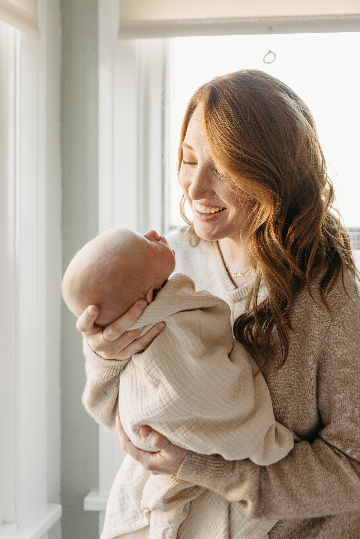 Mom smiles at newborn baby in her arms by window light.