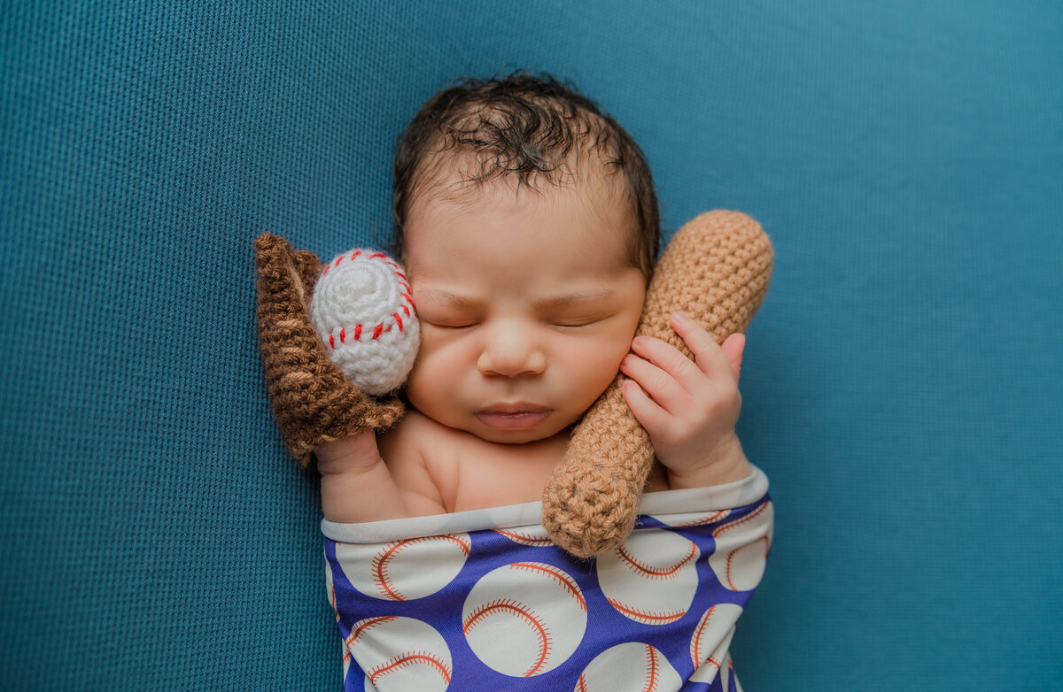 Newborn Photographer, a baby sleeps holding knit baseball mitt, ball, bat, and wrapped in a baseball sheet