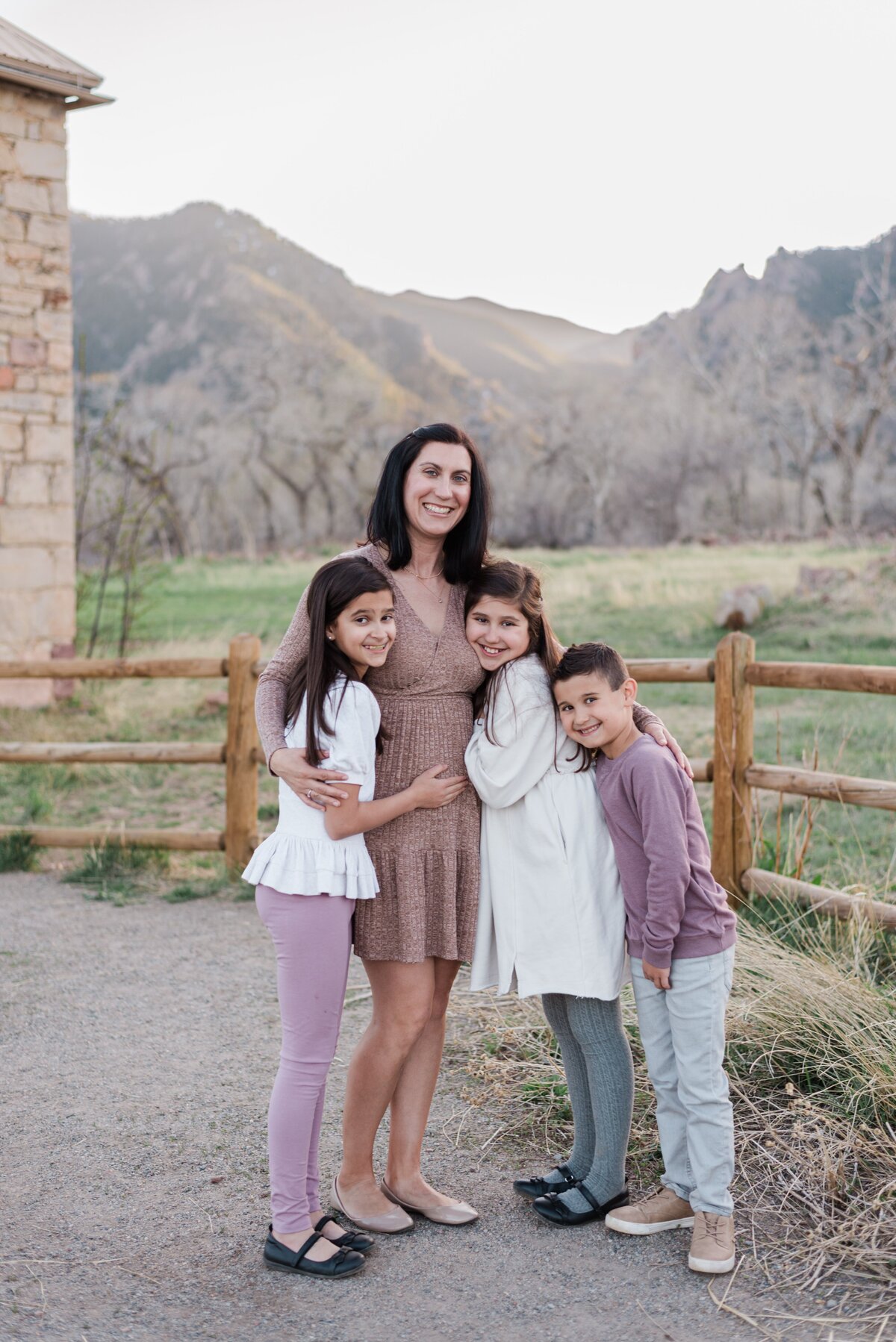 A mom stands with her two daughters and son as they smile toward denver family photographer at south mesa trailhead near the wooden fence along the trail