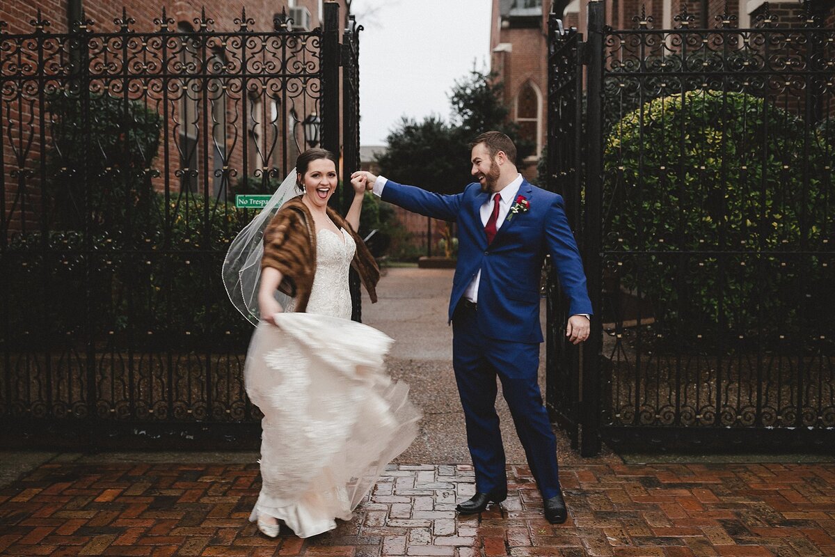 Bride wearing a lace wedding dress, veil and mink stole dances with the groom in a blue suit with a red tie in front of the cast iron gate at a Nashville church.