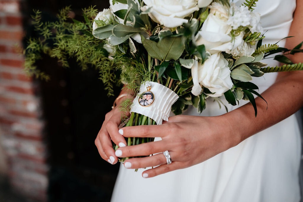 Close-up of a white bridal bouquet from a spring wedding at the Palm Event Center in Pleasanton, CA. The bouquet features a medallion of the bride’s grandparents elegantly wrapped around the flower stems,.
