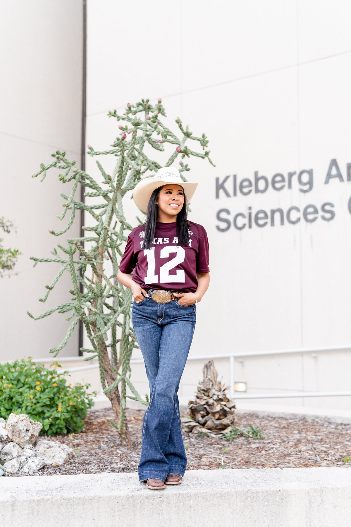 Texas A&M senior girl standing on ledge in front of Kleberg while wearing maroon jersey and jeans with cowboy hat