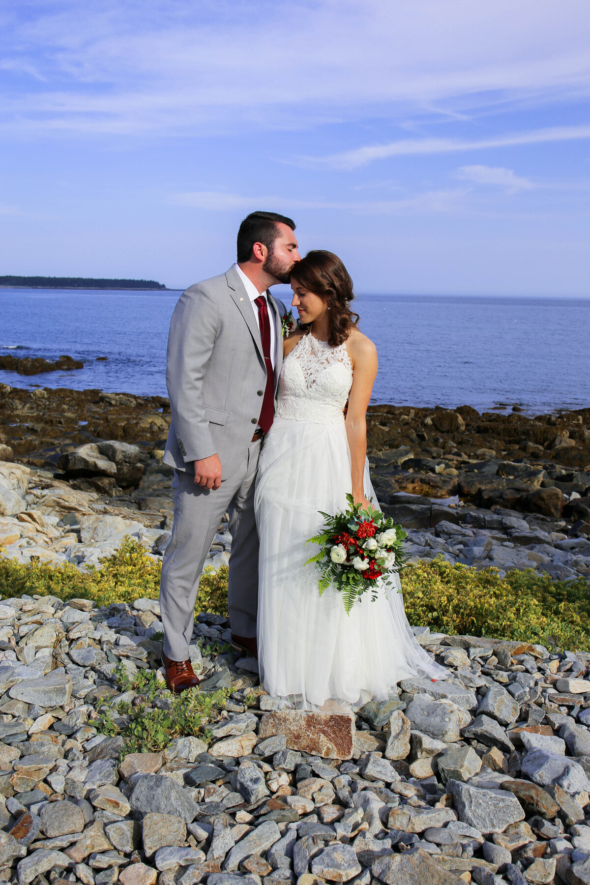Bride and Groom standing on rocky beach, Groom kissing Bride's head | Bar Harbor Wedding Photography