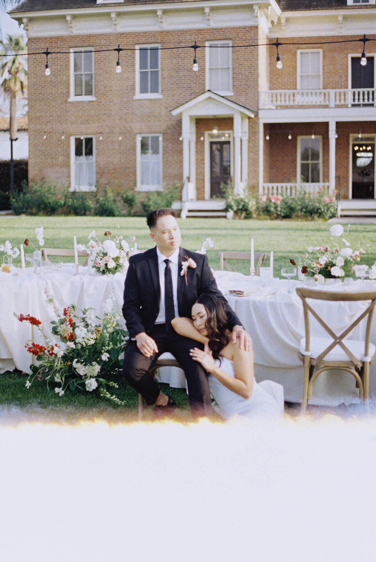 A bride in a white dress rests her head on a groom's knee.