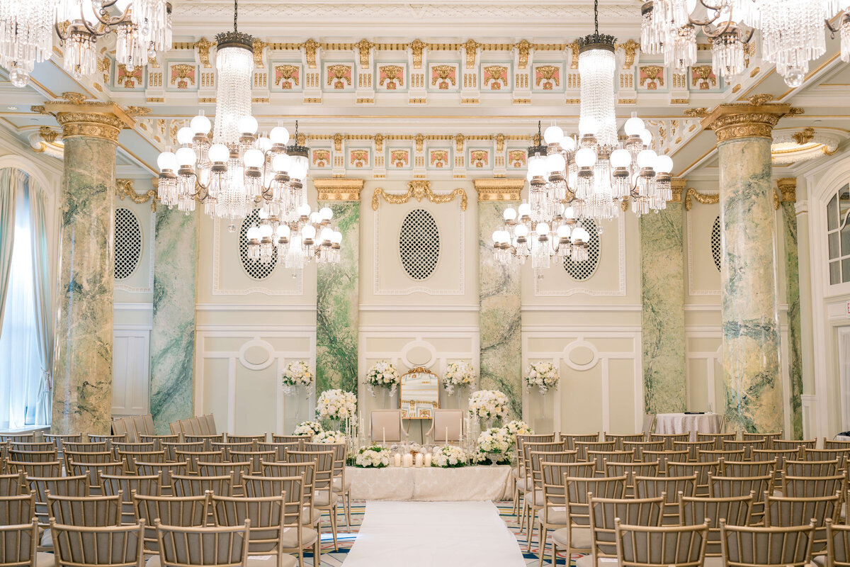 A grand, elegant wedding venue with rows of beige chairs facing an ornate altar. The room features tall marble columns, intricate gold detailing, chandeliers, and floral arrangements, creating a luxurious and sophisticated atmosphere.