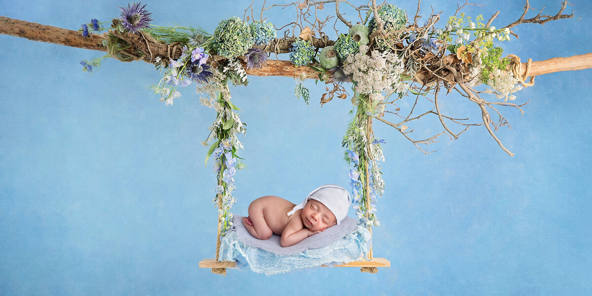 Newborn baby posed on a wooden swing decorated with blue and green florals, sleeping peacefully against a soft blue backdrop.