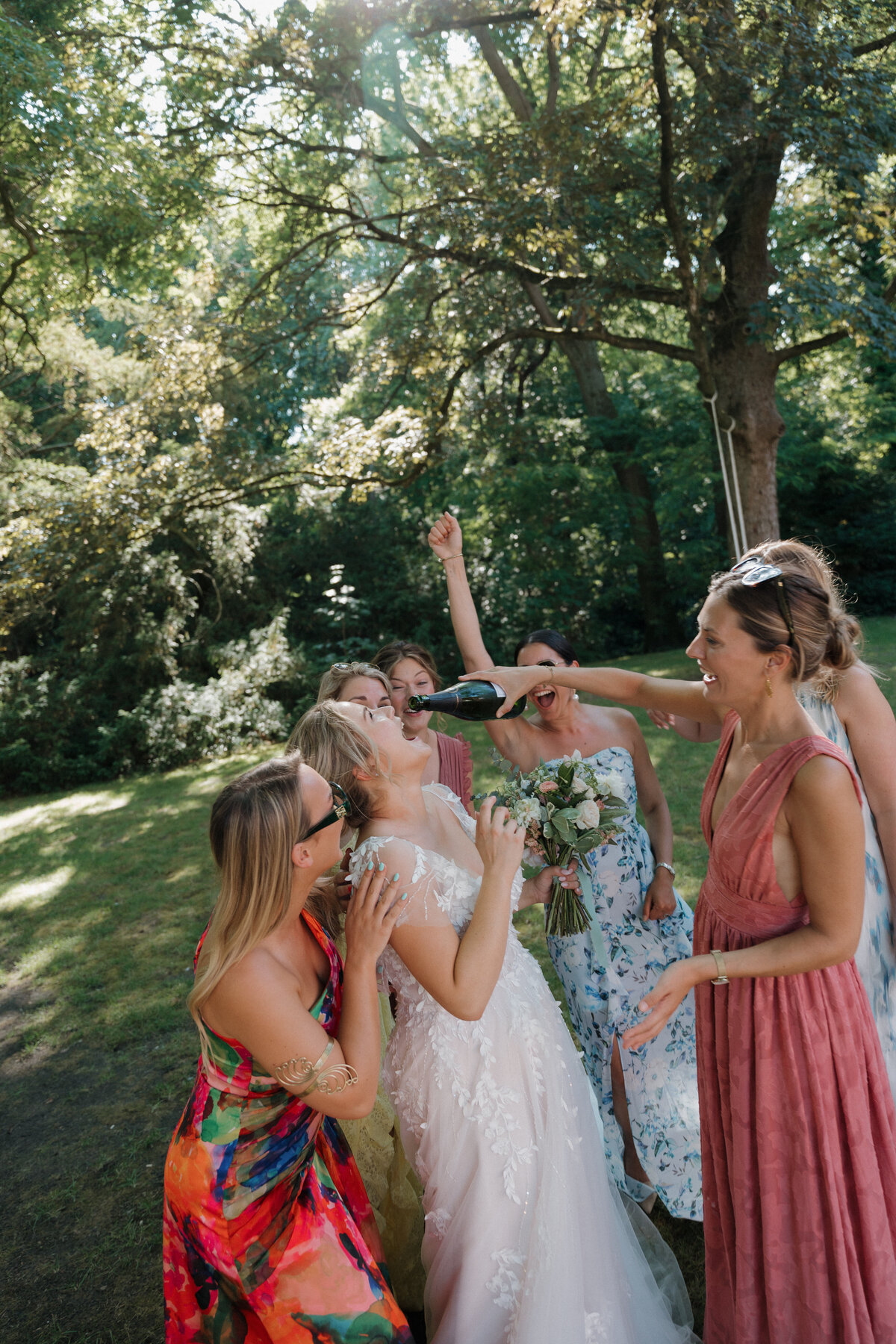 Bruid danst met vriendinnen op het gras onder de bomen en drinkt champagne van de fles.