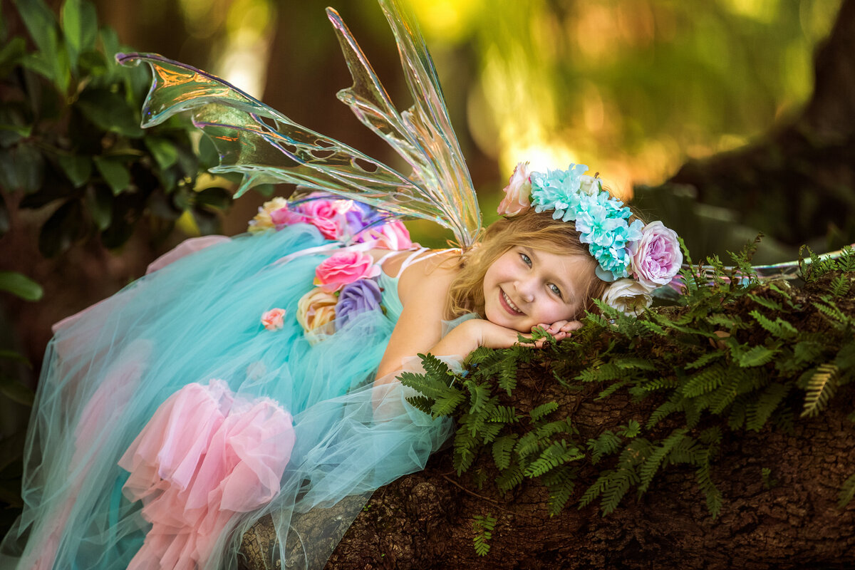 Blonde girl wearing fairy wings by Crone Designs.  She is lying forward on a mossy oak tree branch.  She is wearing an aqua and pink tulle dress with flowers.  She is smiling and looking at the camera.