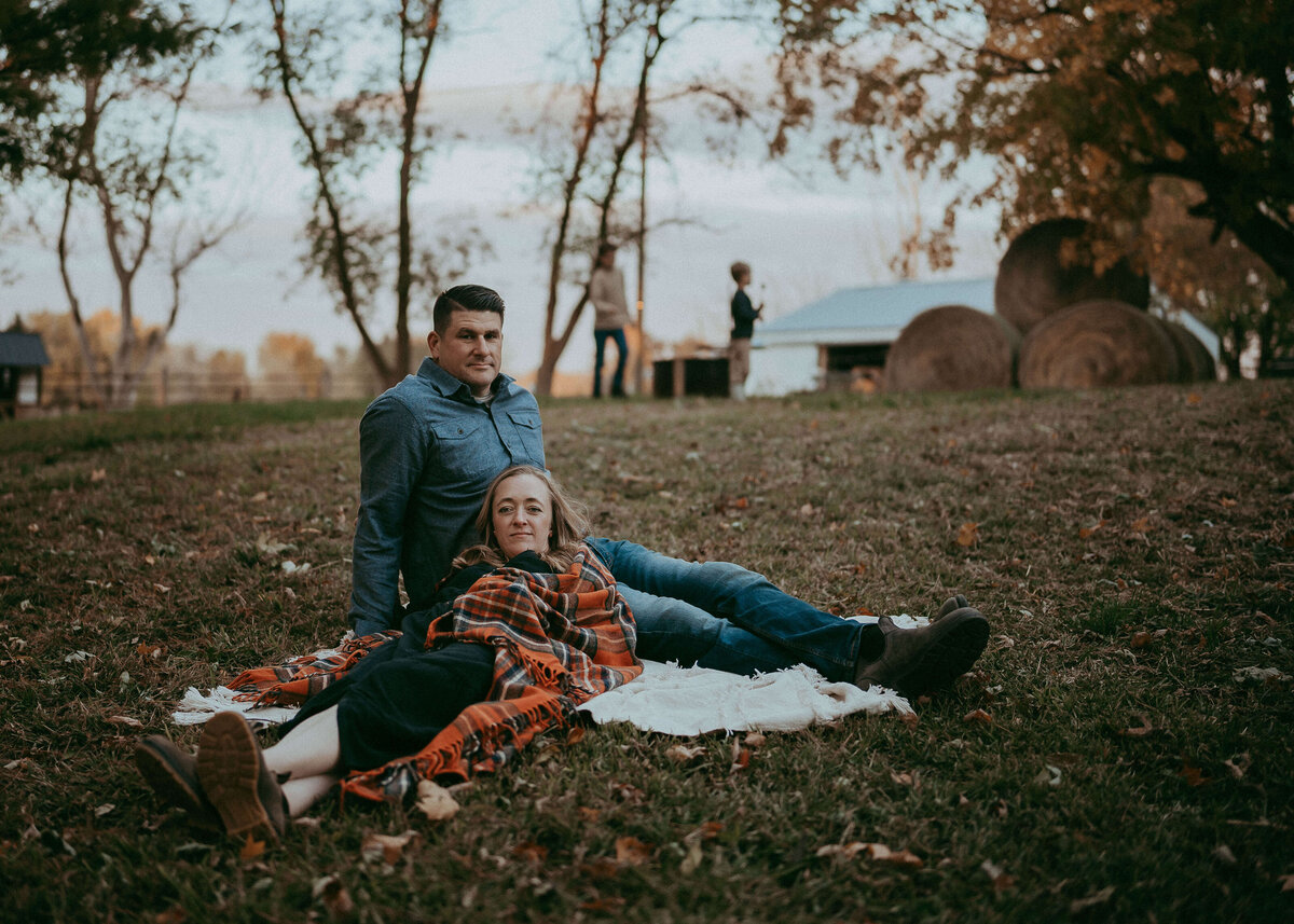 A fine art portrait of a husband and wife sitting on a blanket on their family farm while looking directly at the camera with peaceful, stoic expressions.