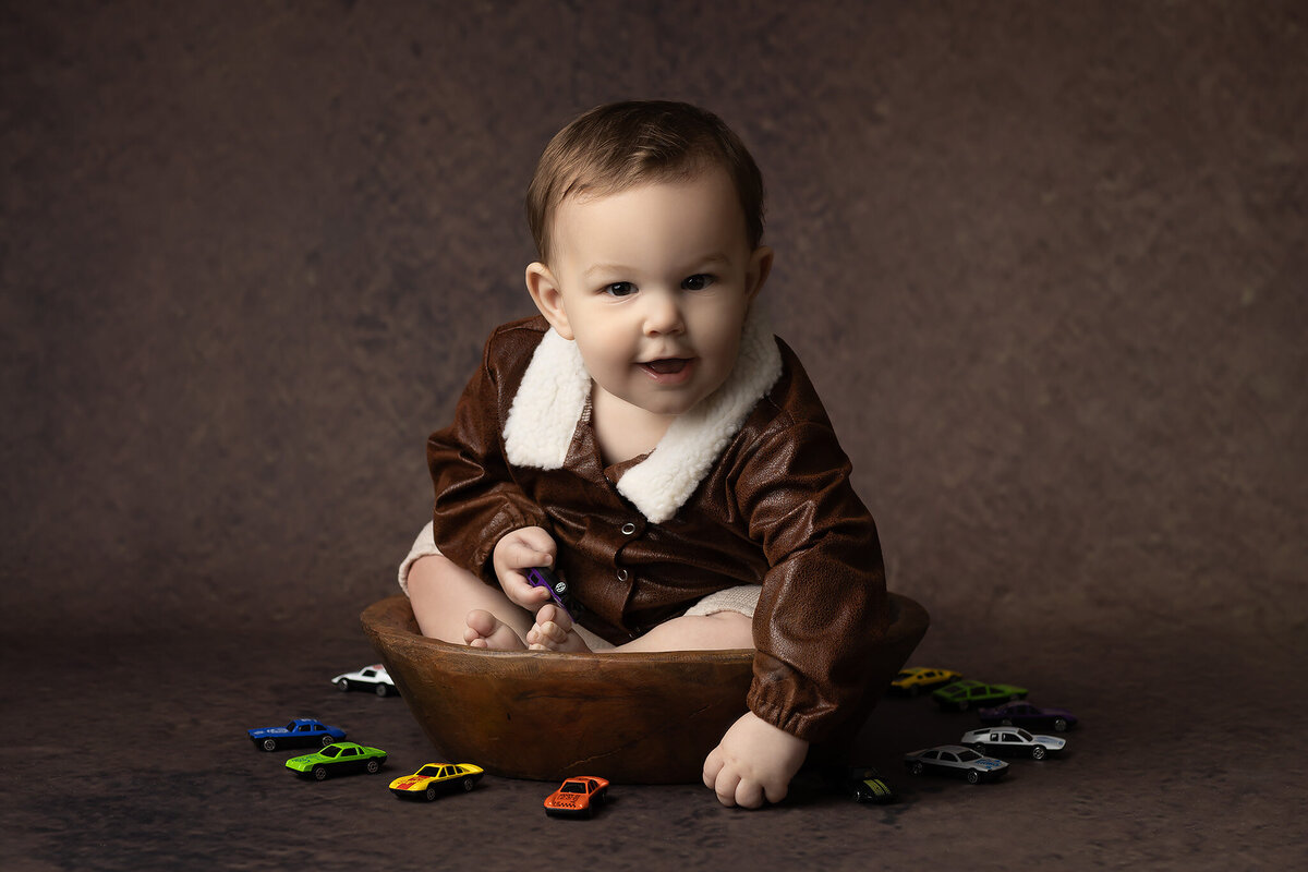Smiling toddler in a faux leather aviator jacket, holding a purple toy car while seated in a wooden bowl surrounded by colorful toy cars, captured by a DFW milestone photographer.