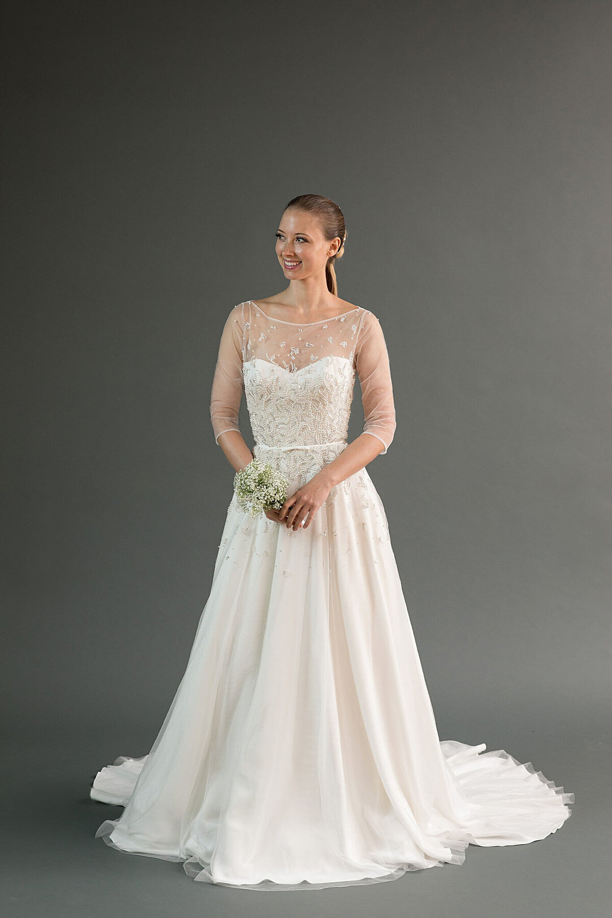 The Rei bridal style is a crystal and pearl beaded wedding dress with pockets, an illusion neckline, and sheer three-quarter sleeves.