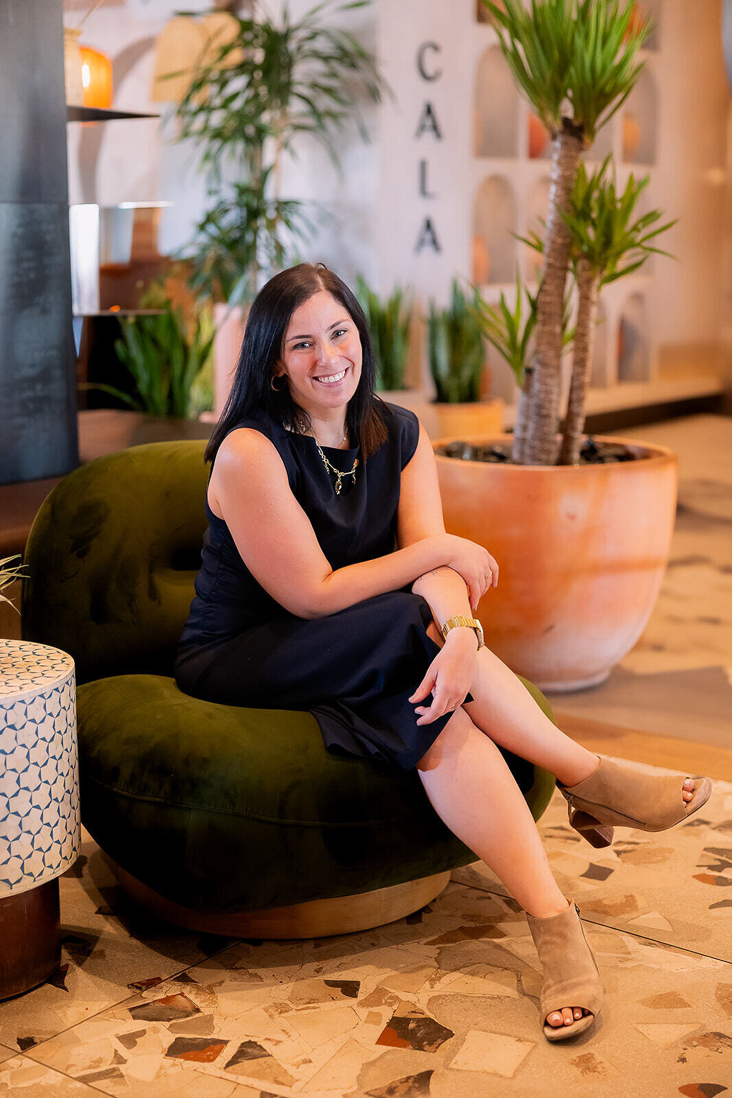 Becky in a black dress sitting with her legs crossed while smiling