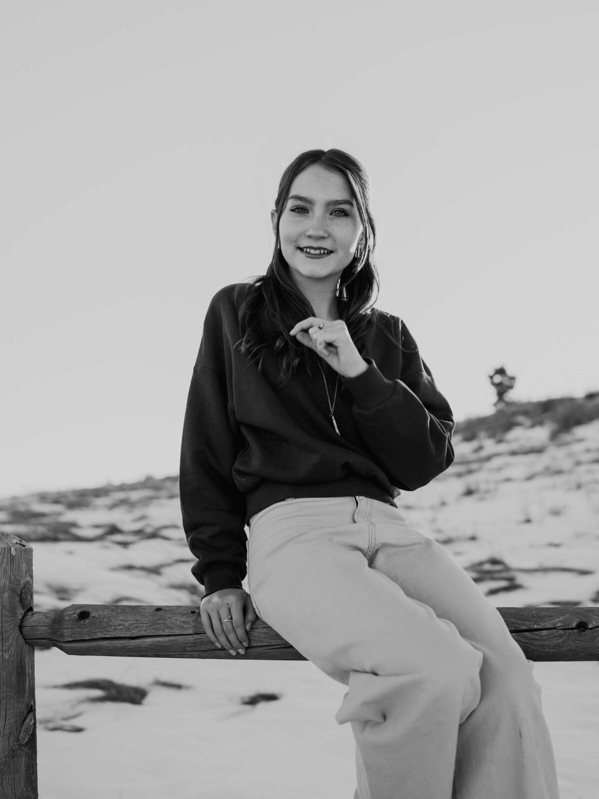 A beautiful close-up, black and white portrait of a teenage girl that was smiling as she holds onto her favorite necklace while sitting on the run of an old farm fence.