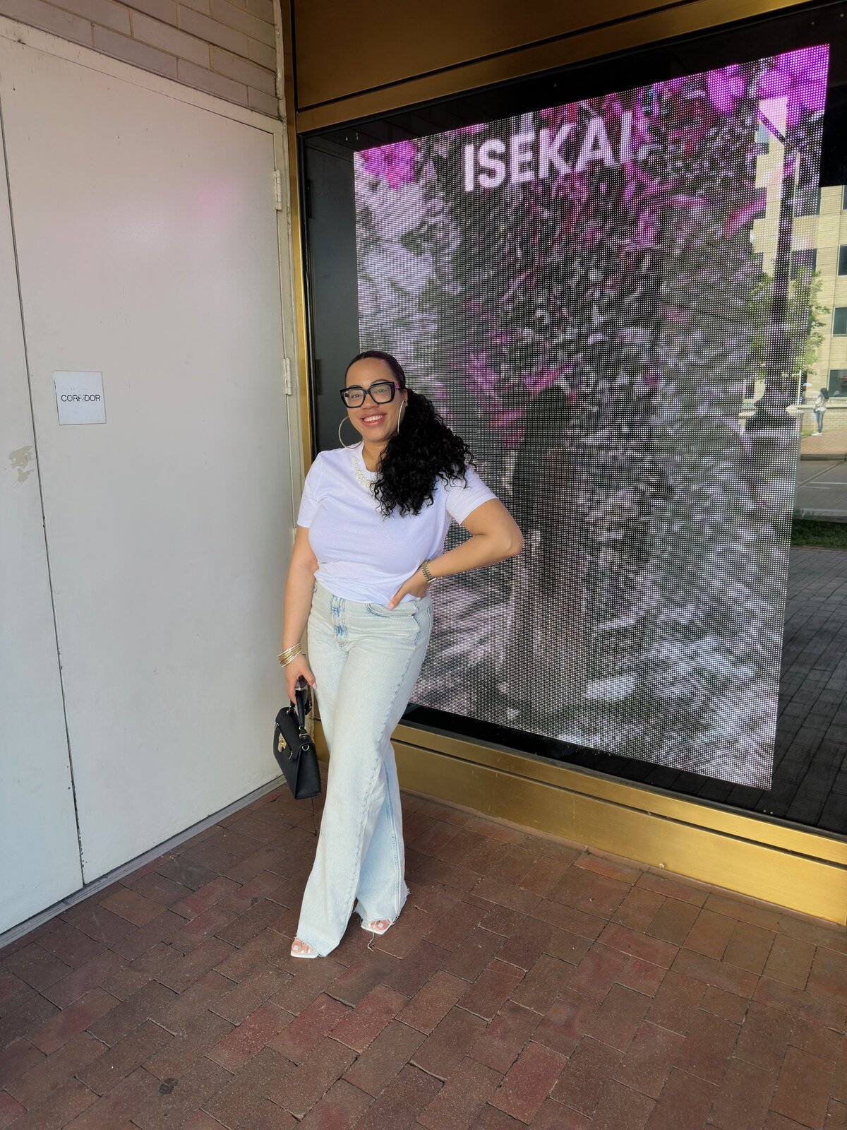 Katrina stands confidently in a white tee and wide-leg jeans, smiling in front of a modern storefront with a digital art display. She holds a black handbag and wears heels, embodying polished, approachable style. 