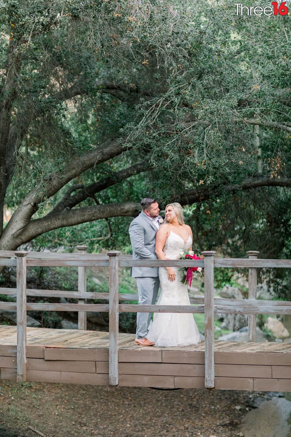 Bride looks back at her Groom as he holds her from behind while standing on a bridge over a small creek