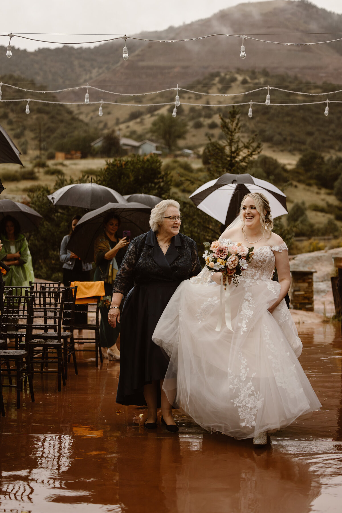 A smiling bride walks arm in arm with her mother down a rain-soaked outdoor aisle under umbrellas, surrounded by wedding guests with mountains in the background.