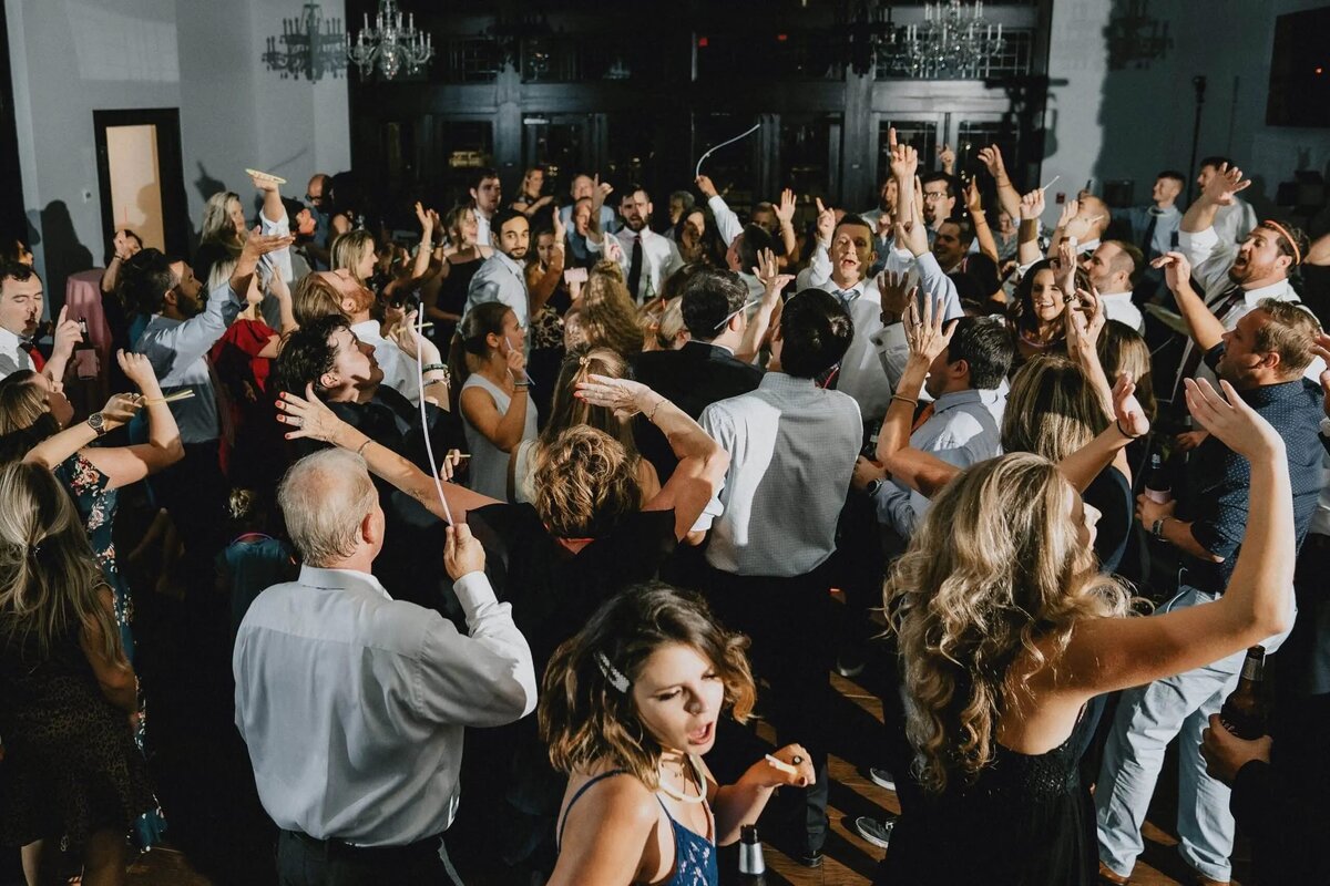 A lively wedding reception dance floor, with guests raising their arms and dancing