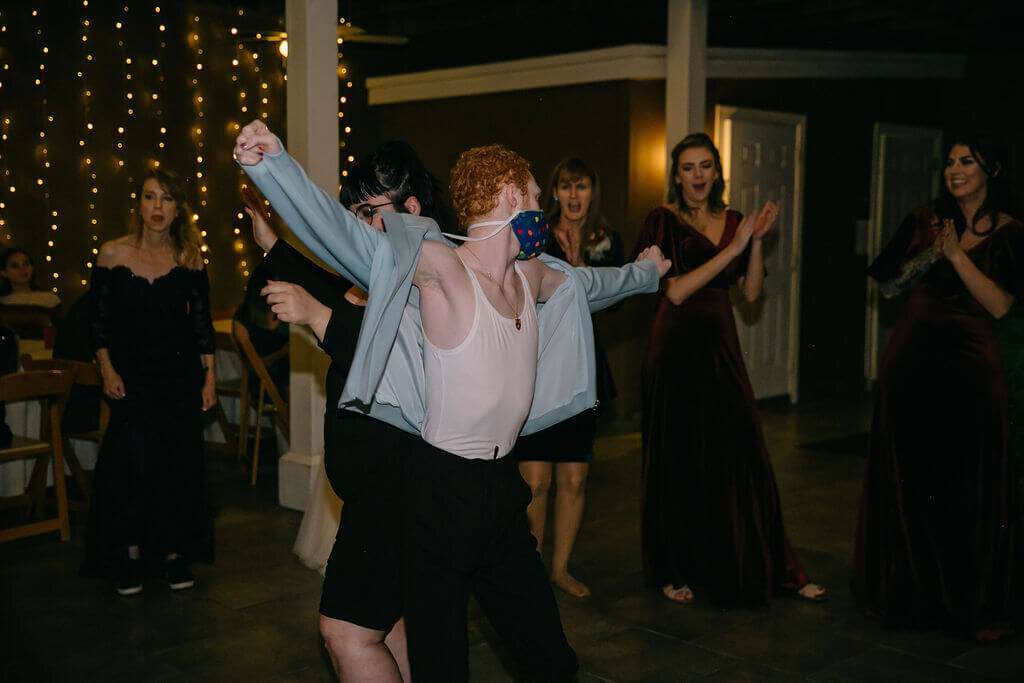 A group of people in formal attire dance. In the center, a person with red hair in a tank top and open jacket dances energetically, wearing a colorful face mask. Twinkling string lights are in the background.