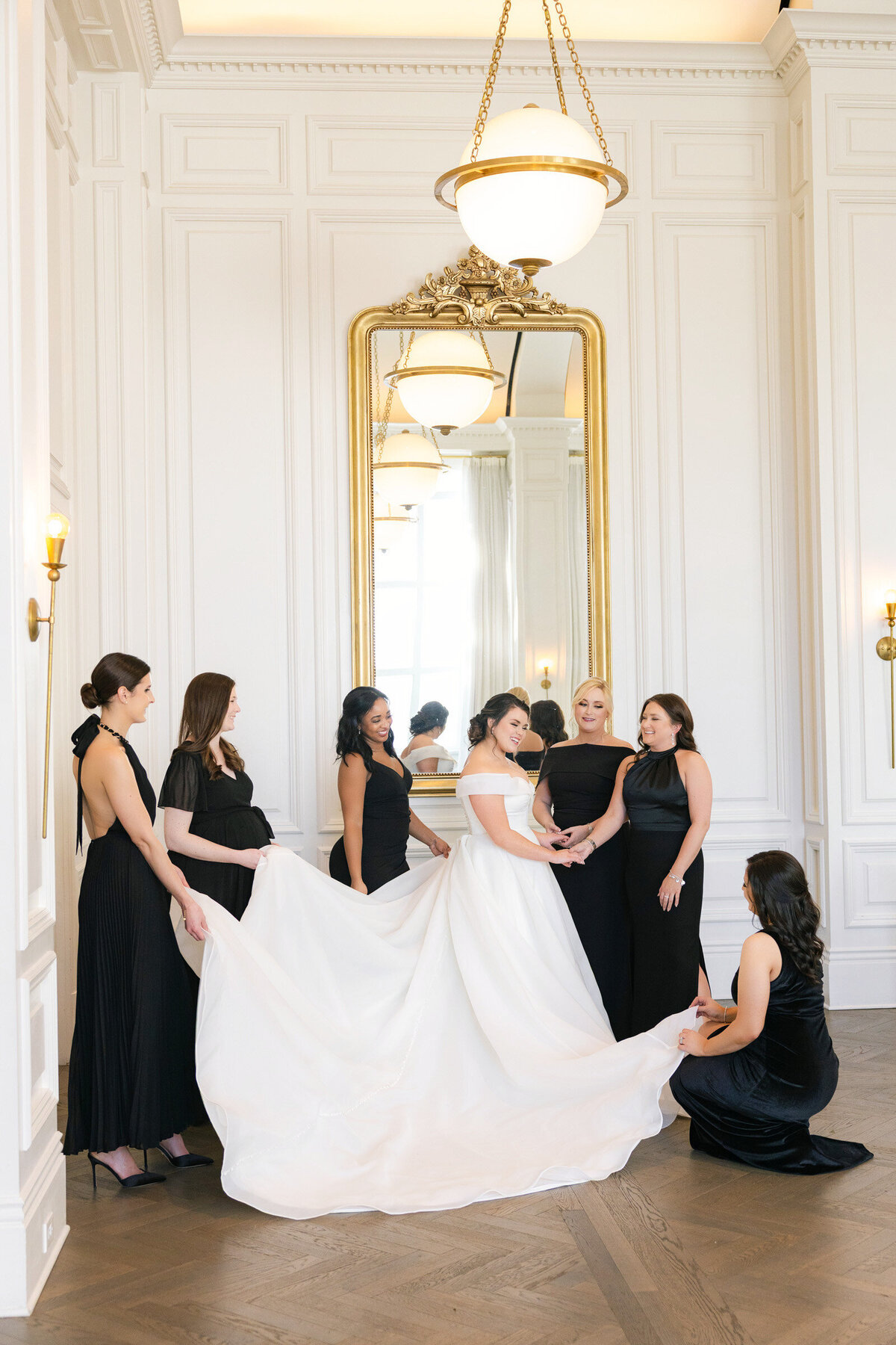 bride surrounded by bridesmaids in black dresses with a large gold-framed mirror behind them in the Governor’s Room at The Adolphus in Dallas, capturing an elegant and stylish wedding portrait.