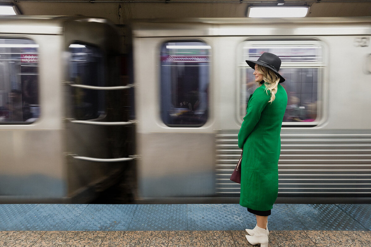 blonde woman in long green coat, standing on the Chicago L platform as train speeds by.