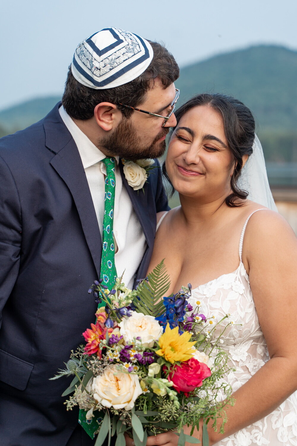 Jewish groom wearing a yarmulke or kippah kissing his bride on the cheek as she scrunches her face in delight