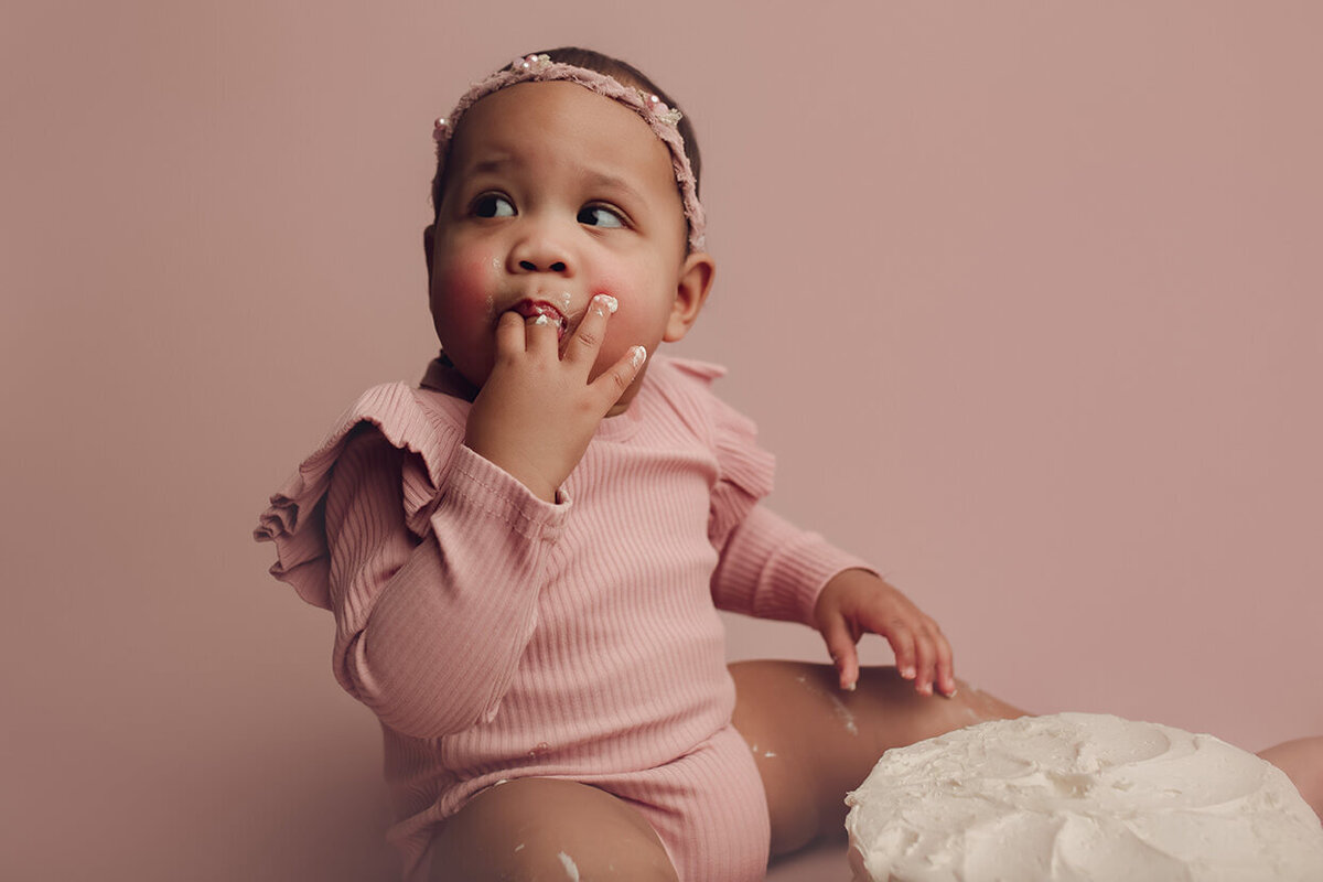 a one year old girl licking cake off her fingers during her cake smash session in rochester ny