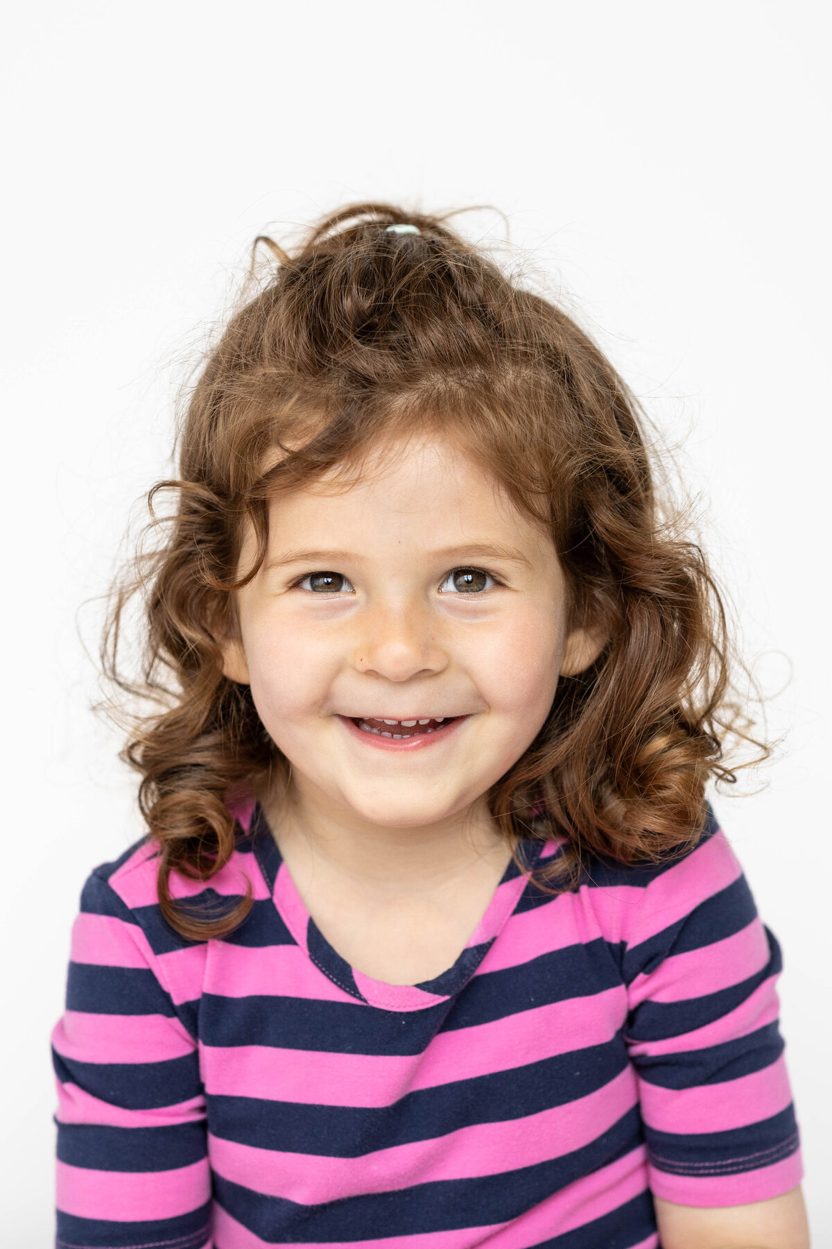 Toddler girl smiling for school photo wearing a purple and navy stripped shirt in front of a white background
