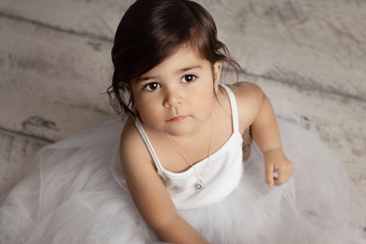 3 year old girl wearing a white dess sitting on a white wooden backdrop  during a child portrait session