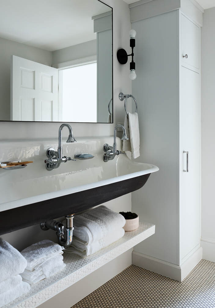 Explore the shared kids bath in a historic Hingham home, featuring a trough sink, tall storage cabinet, open shelf below, penny tile, and a simple, utilitarian design.