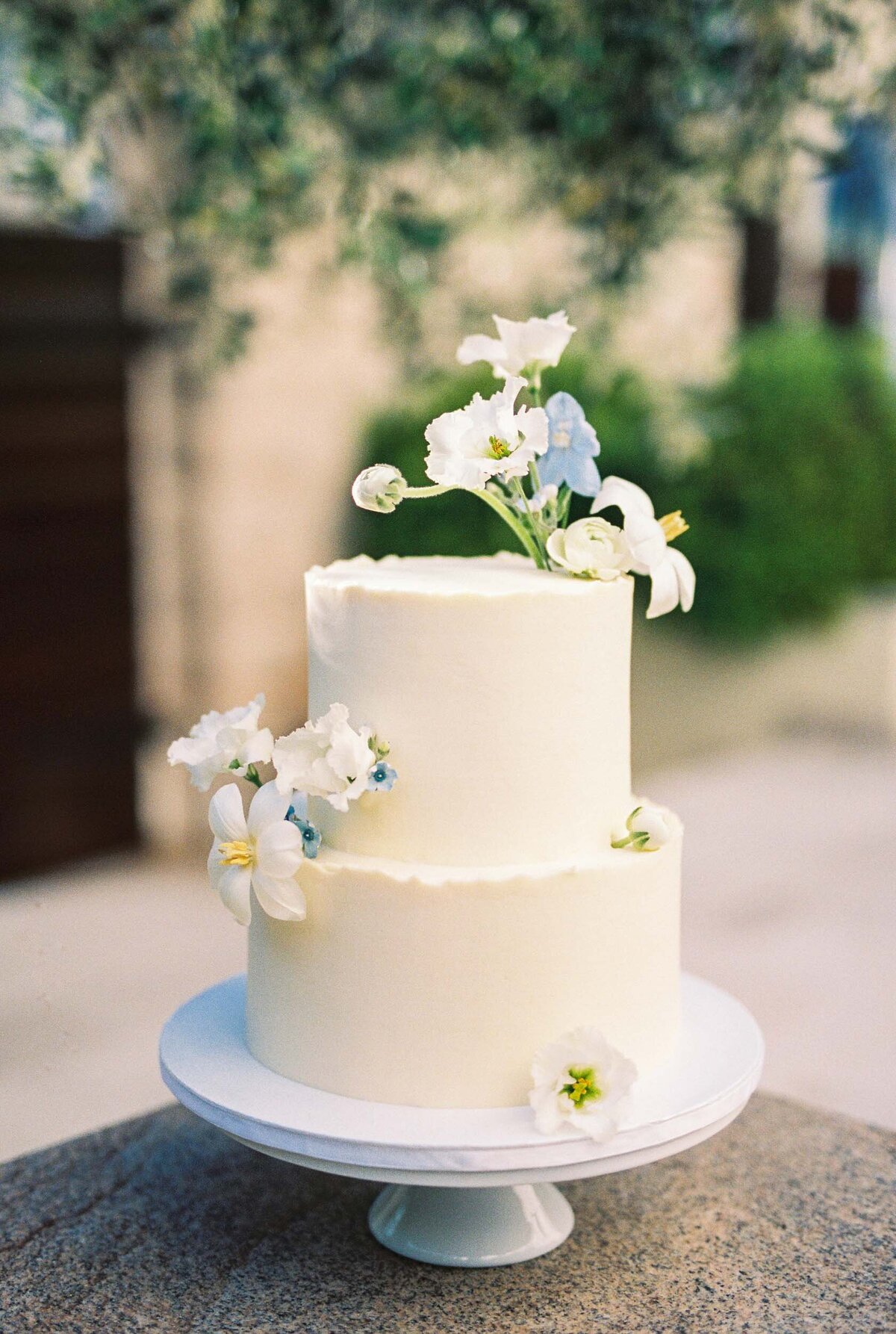 Elegant two-tier white wedding cake adorned with delicate white and blue flowers, photographed in natural light